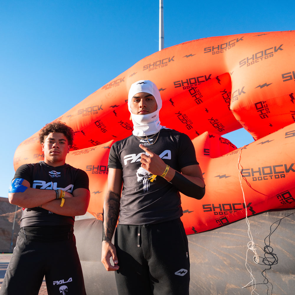 Two 7v7 football athletes standing in front of a large inflatable with 'Shock Doctor' branding. Wearing Shock Doctor White "Sheisty" Ski Mask