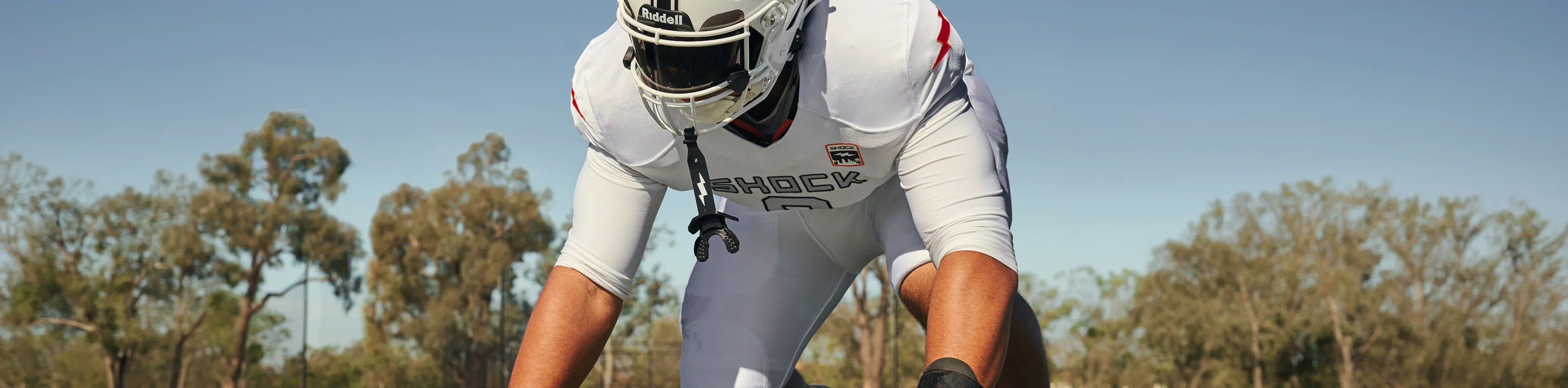 Football player in white Shock uniform with mouthguard hanging, set in a three-point stance