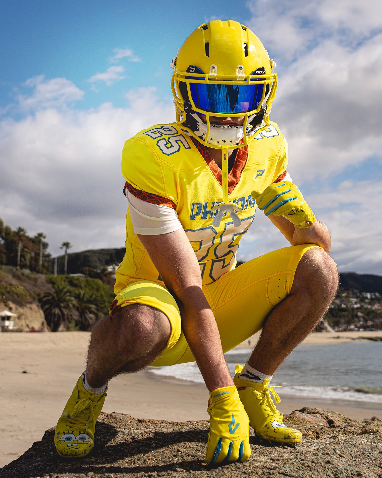 Football player in a yellow Phenom uniform crouching on beach rocks, wearing SpongeBob Quantum Speed 2.0 cleats and gloves