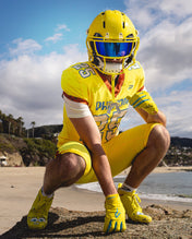 Football player in a yellow Phenom uniform crouching on beach rocks, wearing SpongeBob Quantum Speed 2.0 cleats and gloves