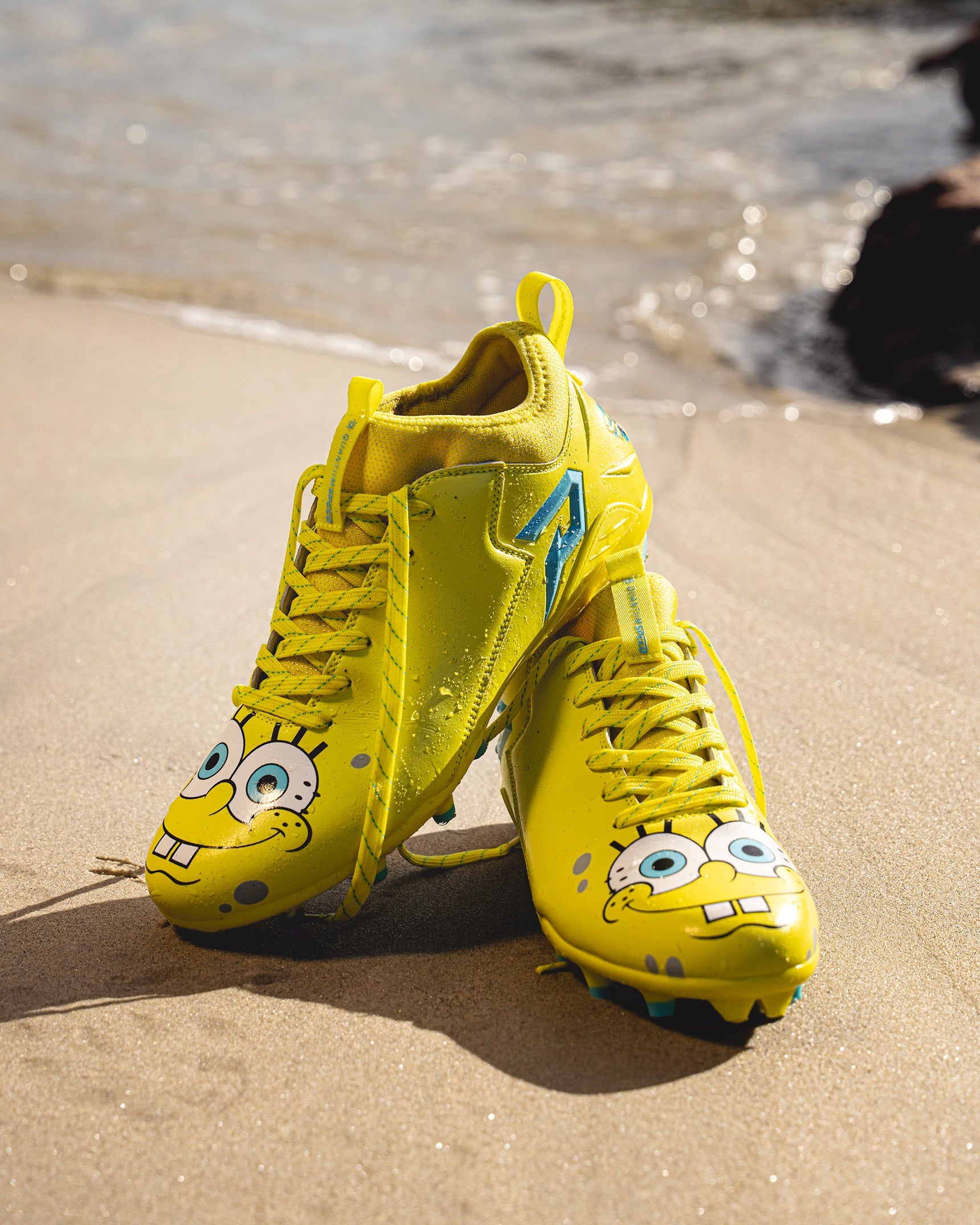 SpongeBob Quantum Speed 2.0 cleats by Phenom Elite propped up on a sandy beach, showing SpongeBob’s face on the toes