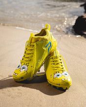 SpongeBob Quantum Speed 2.0 cleats by Phenom Elite propped up on a sandy beach, showing SpongeBob’s face on the toes