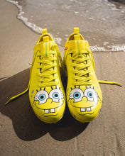 Top-down view of SpongeBob Quantum Speed 2.0 cleats by Phenom Elite on wet sand, with smiling SpongeBob faces on the toes