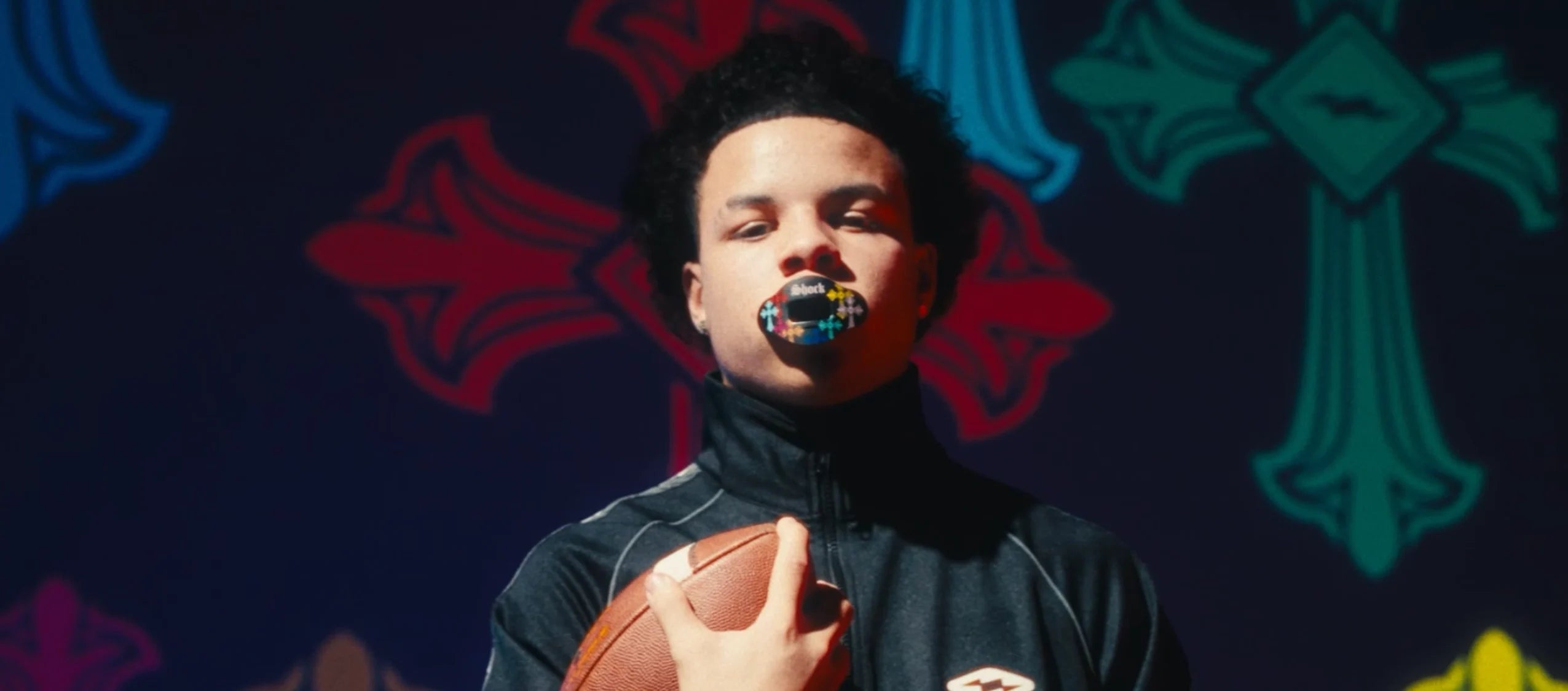 Person holding a football in front of a colorful abstract background wearing Shock Doctor Divine Drip Max AirFlow Lip Guard