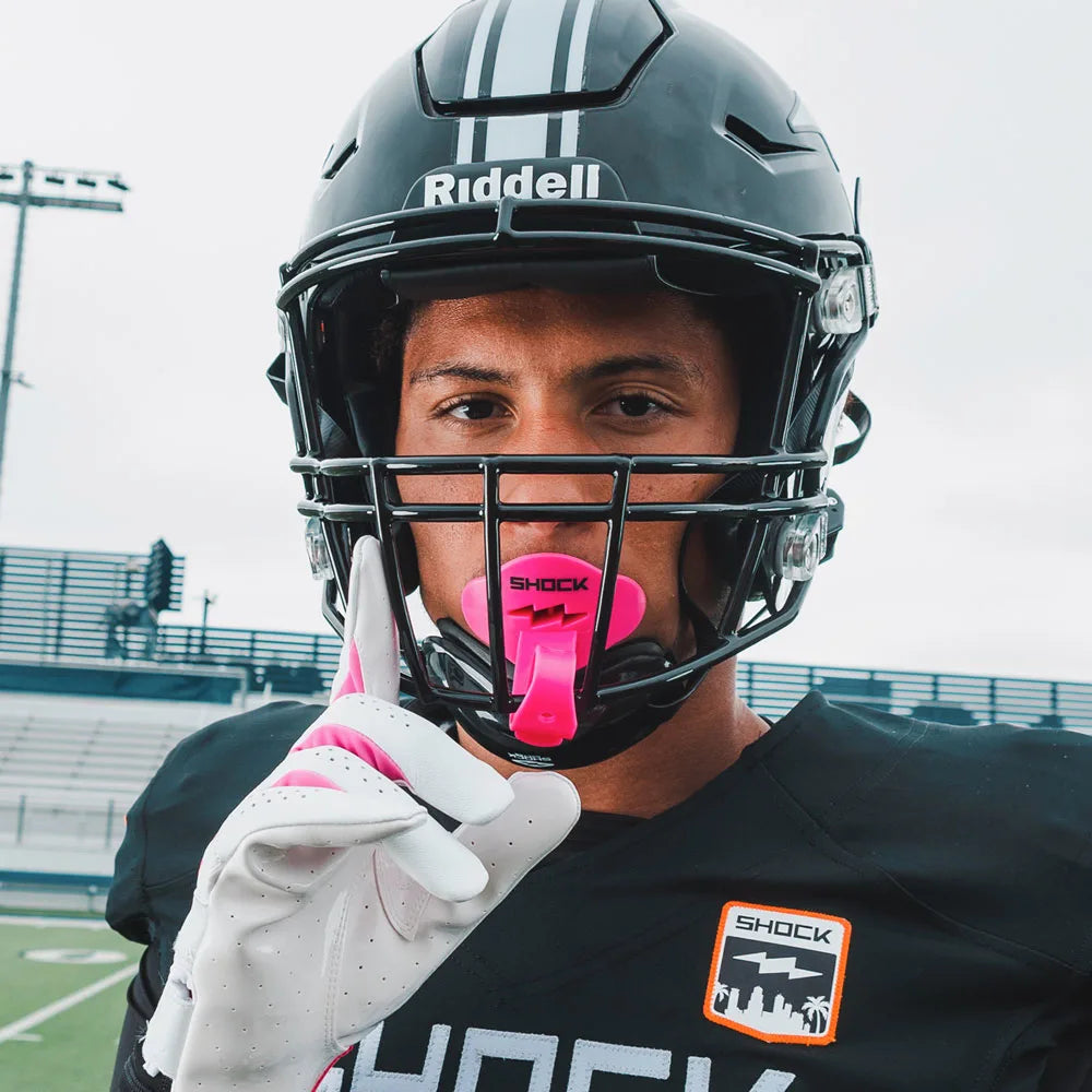 Person wearing a black football helmet with Riddell branding, a pink mouthguard, and white gloves on a football field.