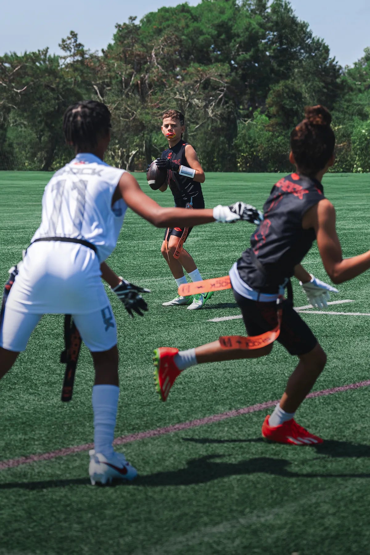 Three male flag football players in action on a field with trees in the background