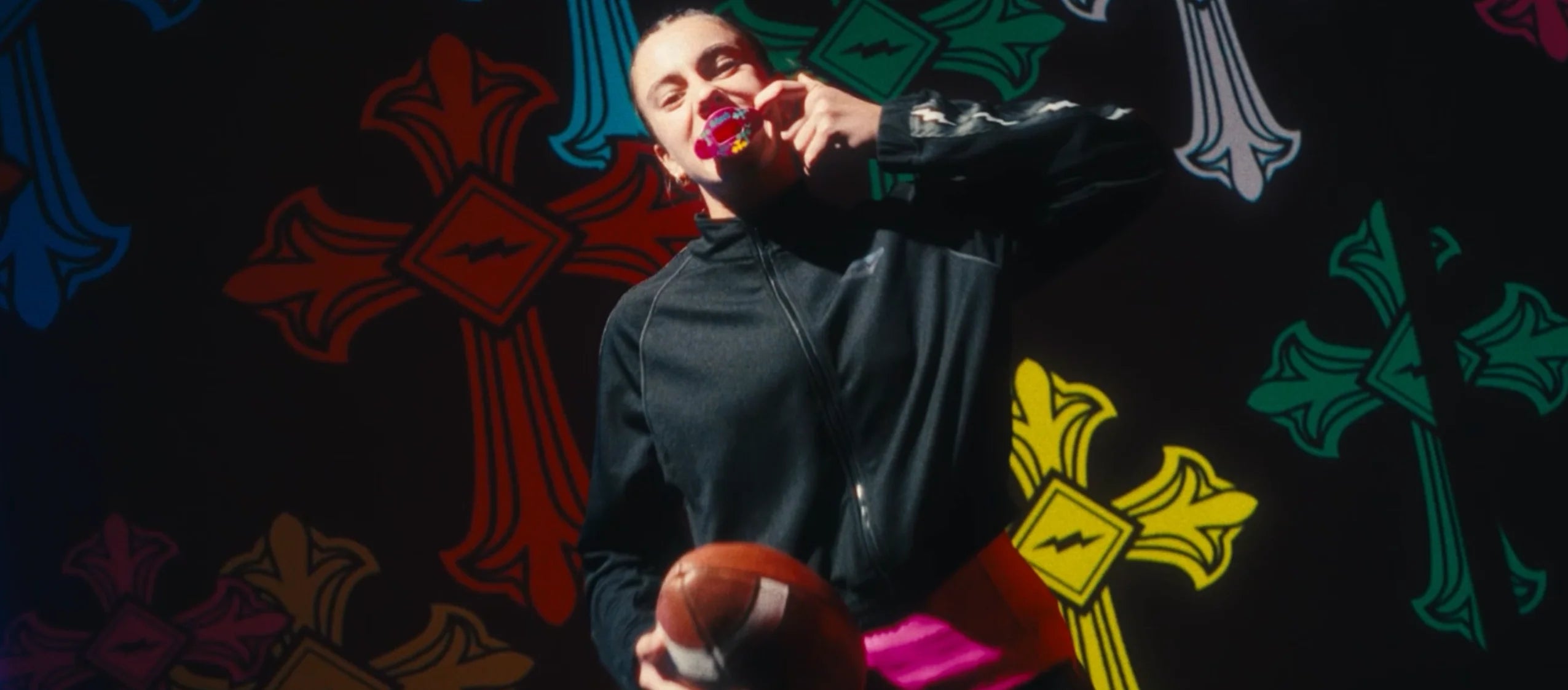 A person in a black zip-up hoodie and black shoes holds a football and wearing a pink lip guard, standing in front of a black background decorated with colorful graffiti-style cross graphics.