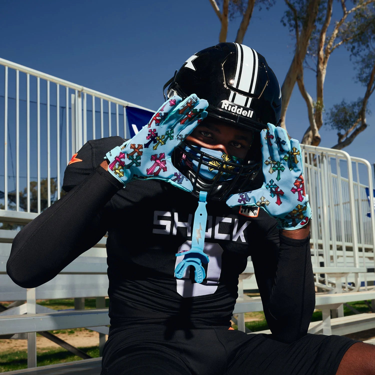 Person wearing a football helmet and gloves with colorful designs, sitting on bleachers.
