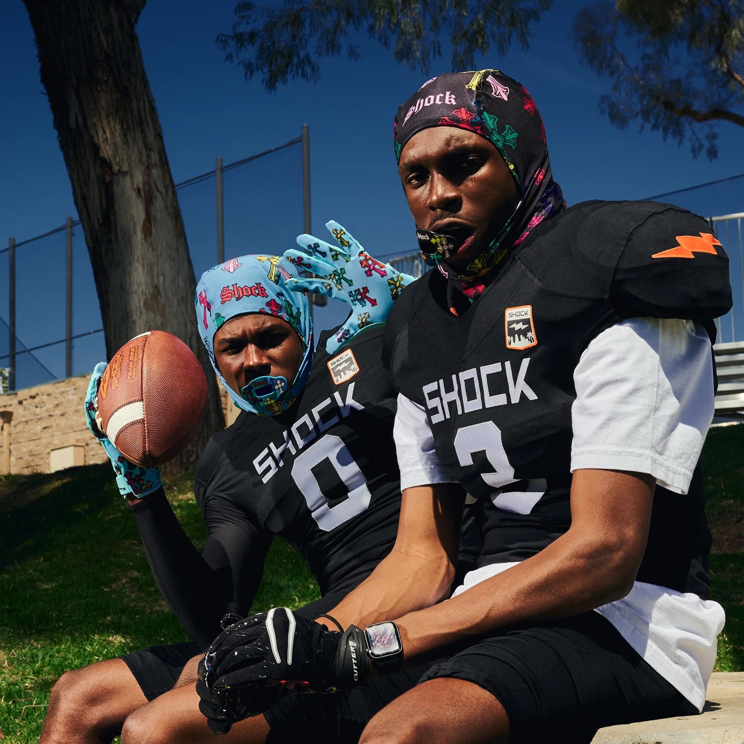 Two football players sitting outdoors wearing black SHOCK jerseys and colorful patterned headwraps, with one holding a football. The scene includes trees, grass, and a fence in the background.