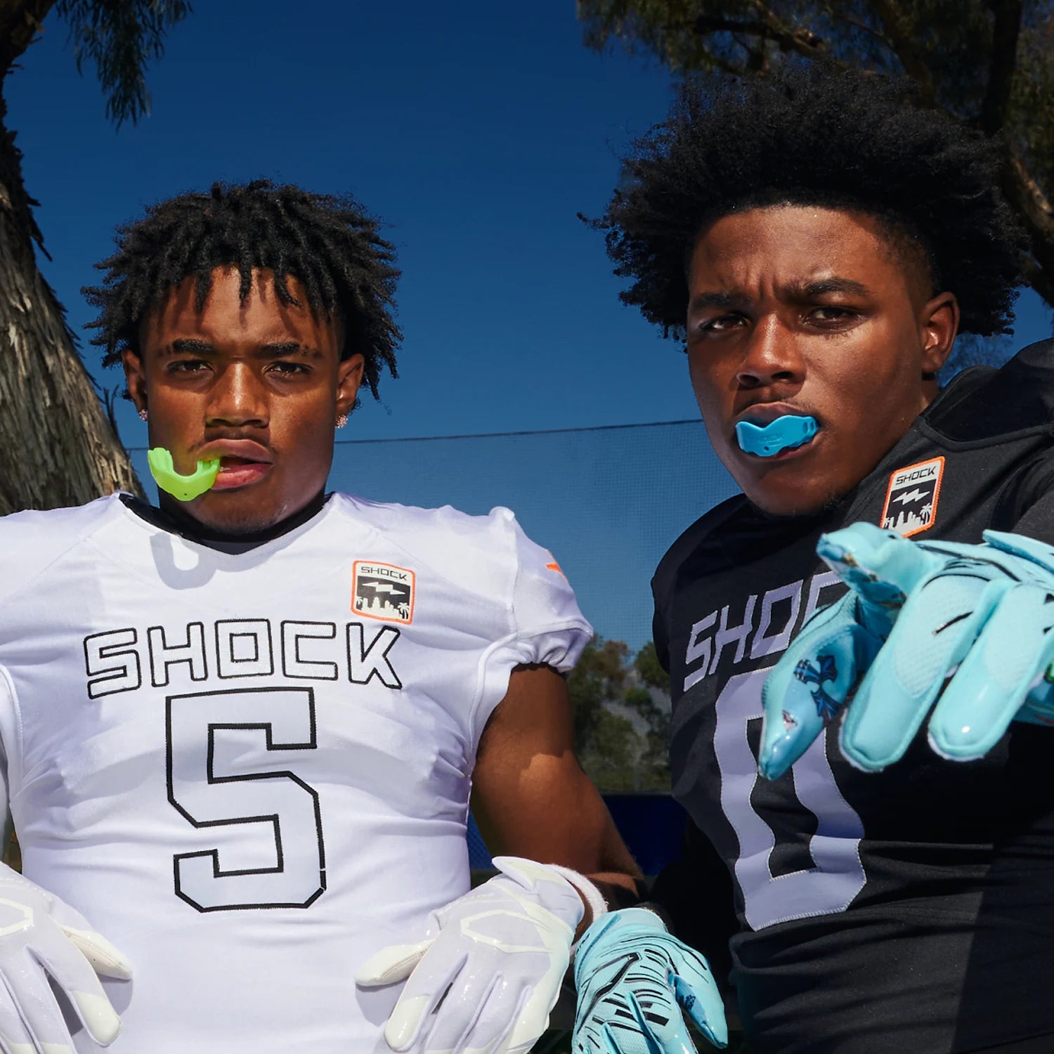 Two young football players outdoors under a blue sky, wearing athletic jerseys and gloves. The player on the left wears a white jersey with the number 5 and the word 'SHOCK' across the chest, while the player on the right wears a black jersey with 'SHO' visible and light blue gloves.