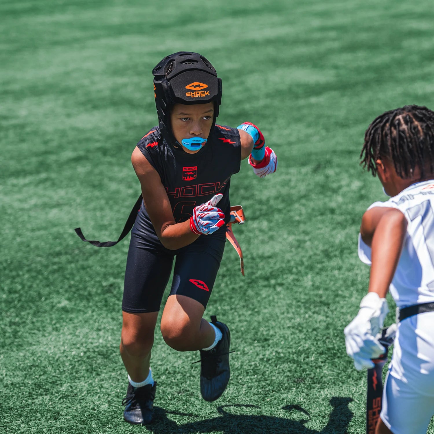 A child athlete wearing a black protective footabll headguard, black athletic shorts, and gloves runs across a green turf field. Another child in a white uniform is partially visible on the right.