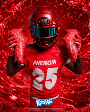 Football player in a red Phenom uniform holding up Kool-Aid Cherry cleats against a red liquid splash