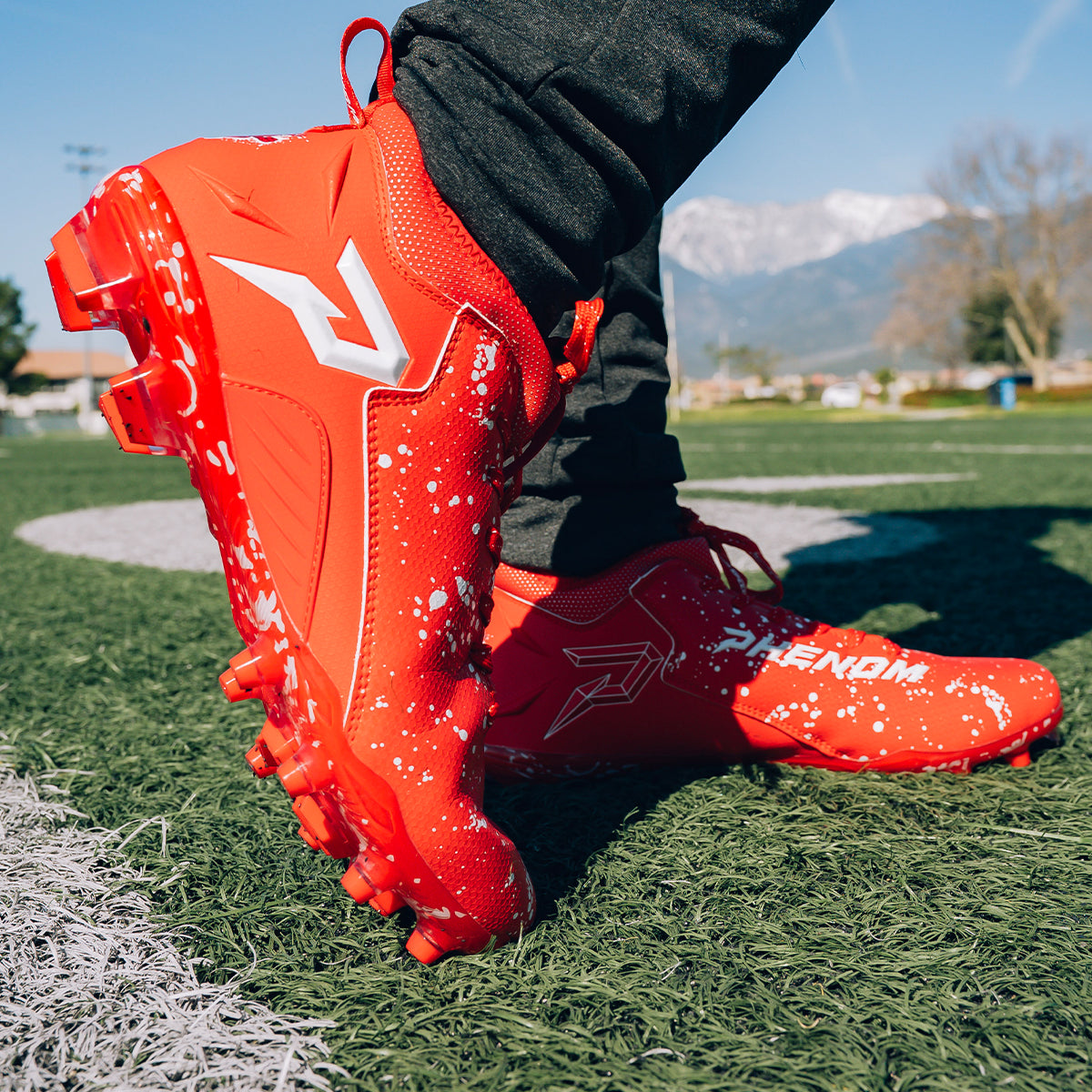 Athlete in Kool-Aid Cherry cleats by Phenom Elite on a turf field, side view with white splatter and P logo, mountains behind