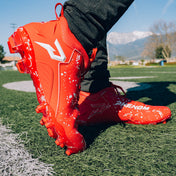 Athlete in Kool-Aid Cherry cleats by Phenom Elite on a turf field, side view with white splatter and P logo, mountains behind
