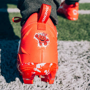 Close-up of a Kool-Aid Cherry cleat by Phenom Elite on turf, showing the Kool-Aid Man heel graphic and PHENOM pull tab