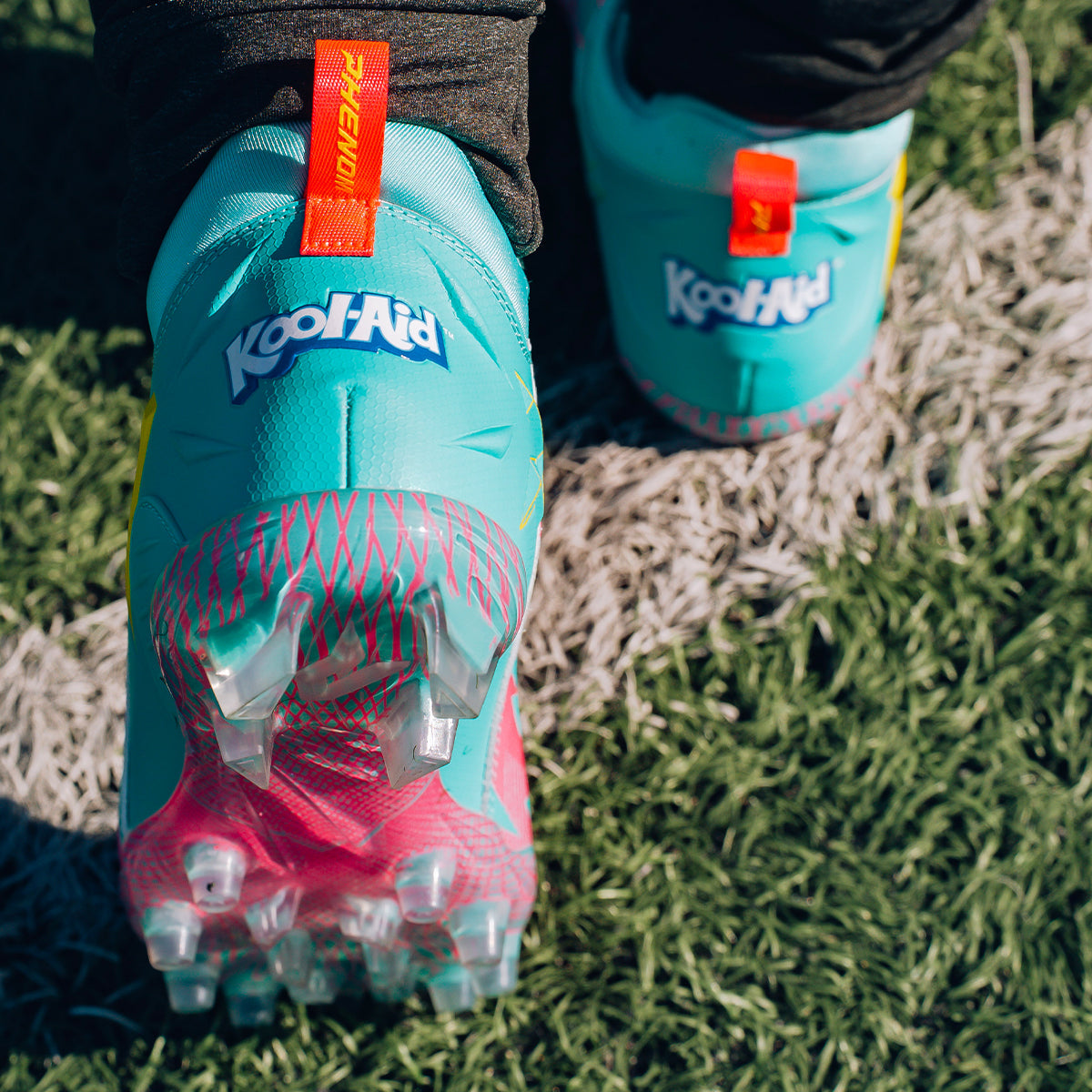 Close-up of Kool-Aid Sharkleberry Fin cleats by Phenom Elite on turf, showing the Kool-Aid heel logo and clear studs