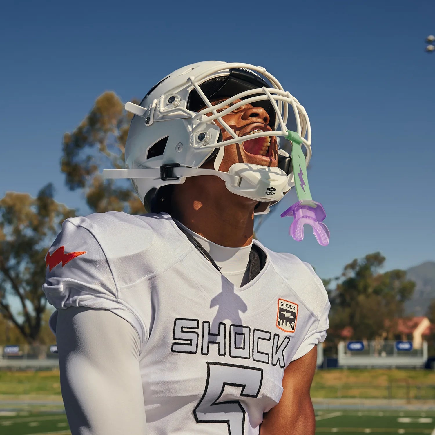 A football player wearing a white jersey with 'SHOCK' and the number 5, along with a white helmet and lipguard in green and purple, standing on a field under a blue sky.