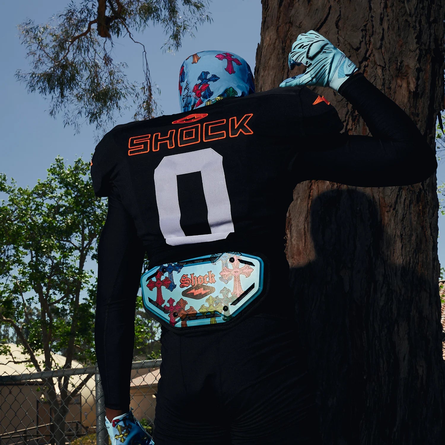 A person wearing a black long-sleeve jersey with the word 'SHOCK' and large white number 0 on the back, standing outdoors against a tree trunk. The jersey has colorful graphic details, including a decorative patch at the waist and gloves, under bright daylight.