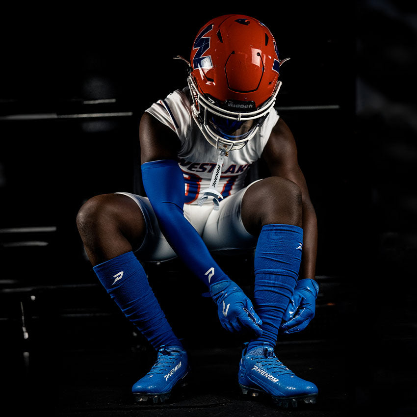 Football player in uniform with helmet and blue gear sitting on a bench.