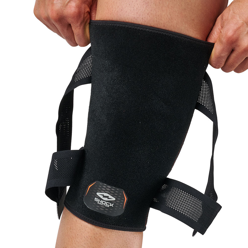 Person pulling the top edge of the Shock Doctor Cross Compression Knee Sleeve into place on their leg, with the unfastened straps hanging loose on either side