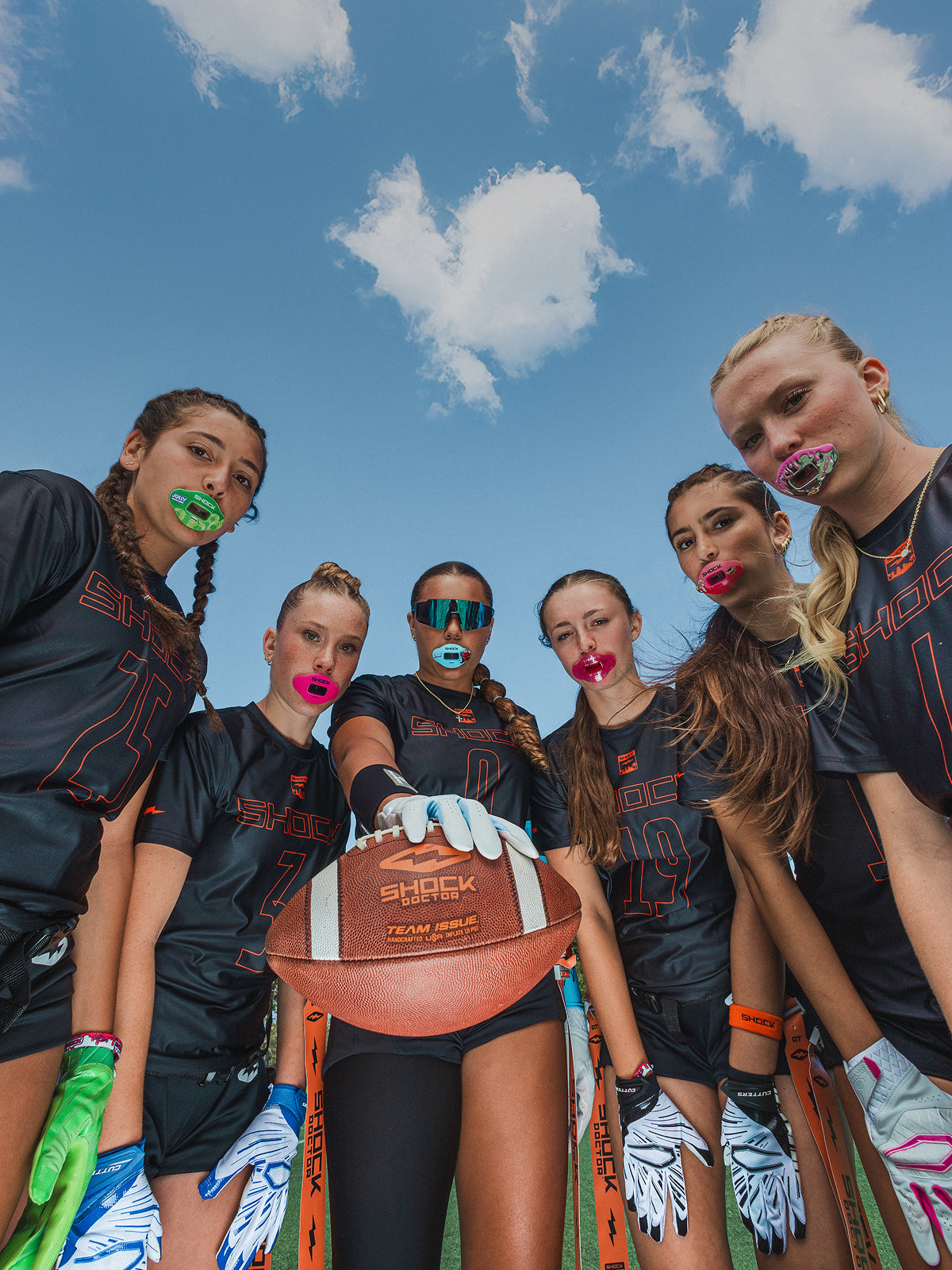 Five women in sports jerseys with colorful football lip guards, against a clear blue sky.