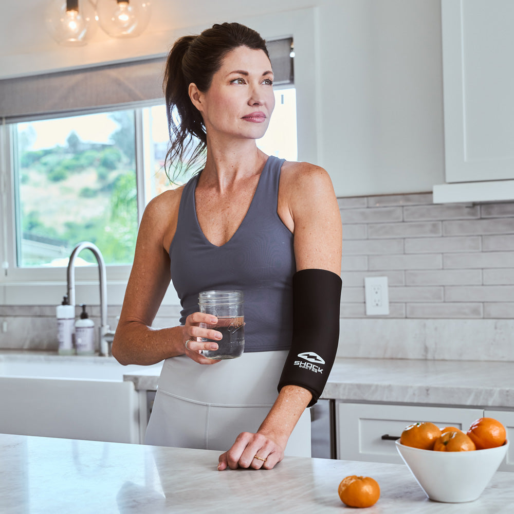Woman holding a glass of water at a kitchen counter, wearing the Shock Doctor Flex Ice Therapy Arm/Elbow Compression Sleeve