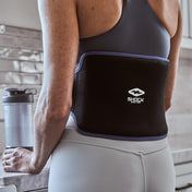 Close-up of a woman’s lower back wearing the black Shock Doctor Flex Ice Therapy Back Compression Wrap in a kitchen
