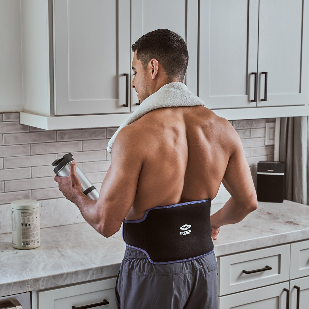 Man in a kitchen holding a shaker bottle, wearing the Shock Doctor Flex Ice Therapy Back Compression Wrap after a workout