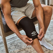 Man seated on a wooden chair pulling the black Shock Doctor Flex Ice Therapy Knee/Thigh Compression Sleeve up over his knee