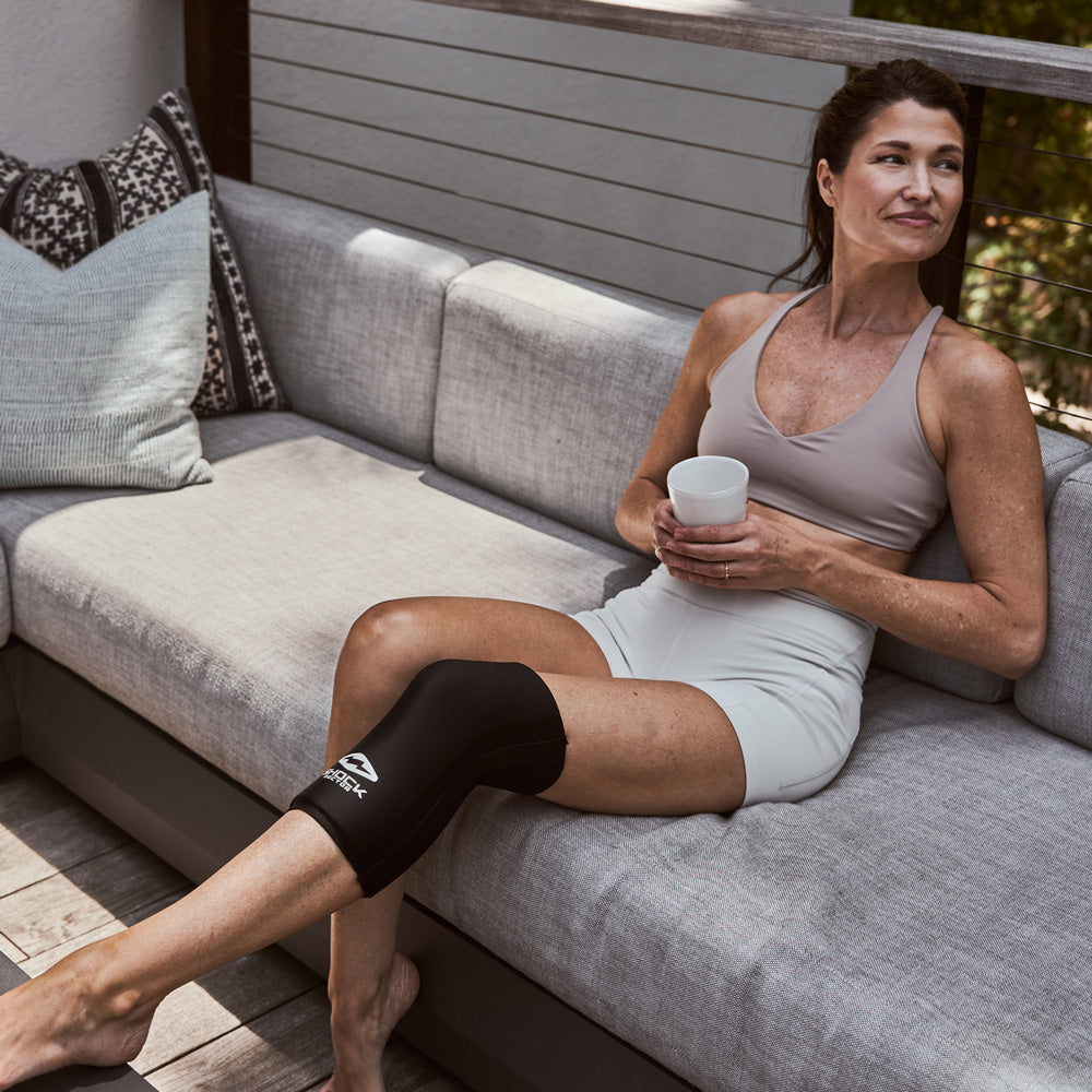Woman relaxing on an outdoor sofa with a coffee mug, wearing the Shock Doctor Flex Ice Therapy Knee/Thigh Compression Sleeve