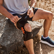 Man seated on a rock on a dirt trail, securing the Shock Doctor NRG Spring Hinge Knee Over Wrap with orange spring hinges