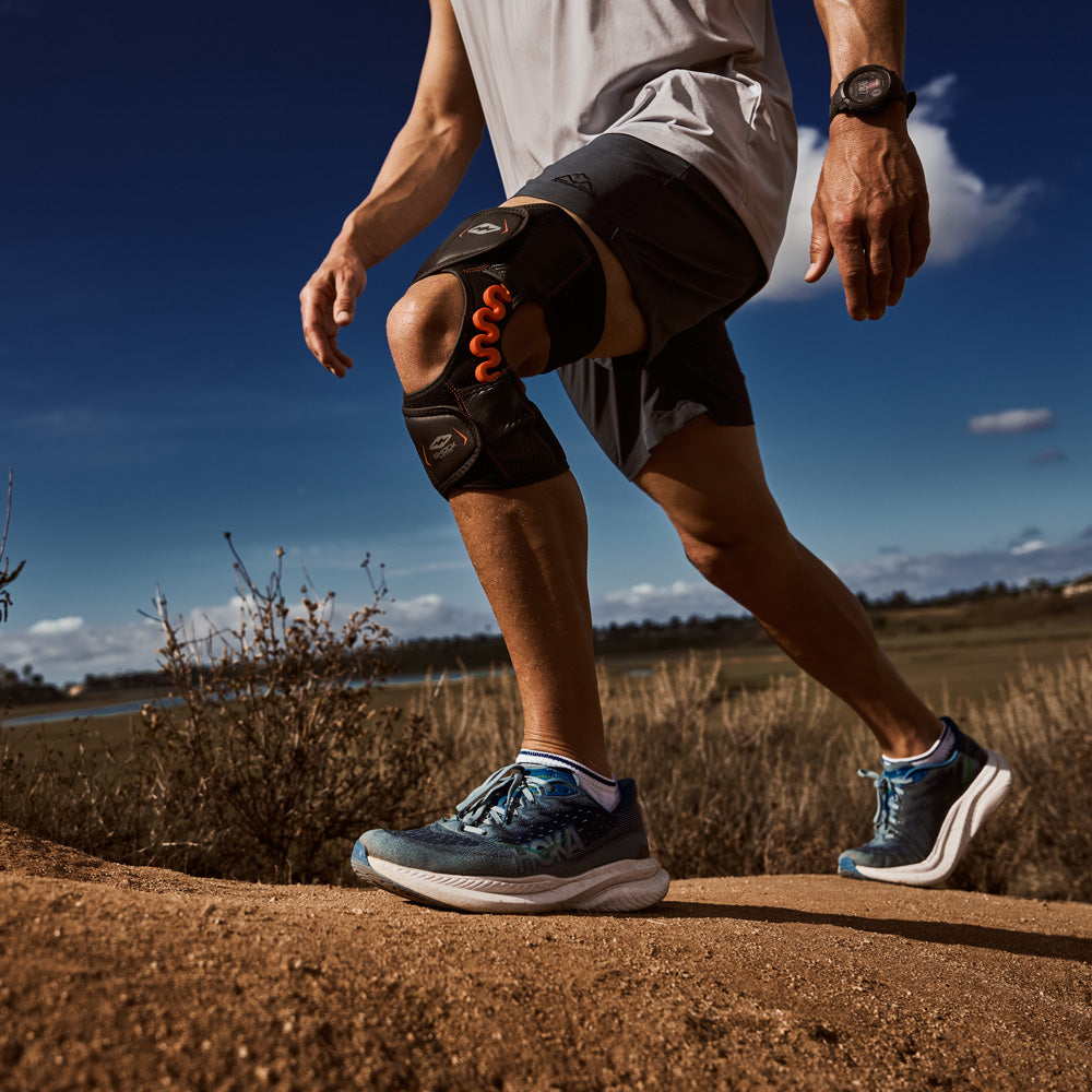 Low-angle view of a man hiking a dirt trail under a blue sky wearing the Shock Doctor NRG Spring Hinge Knee Over Wrap
