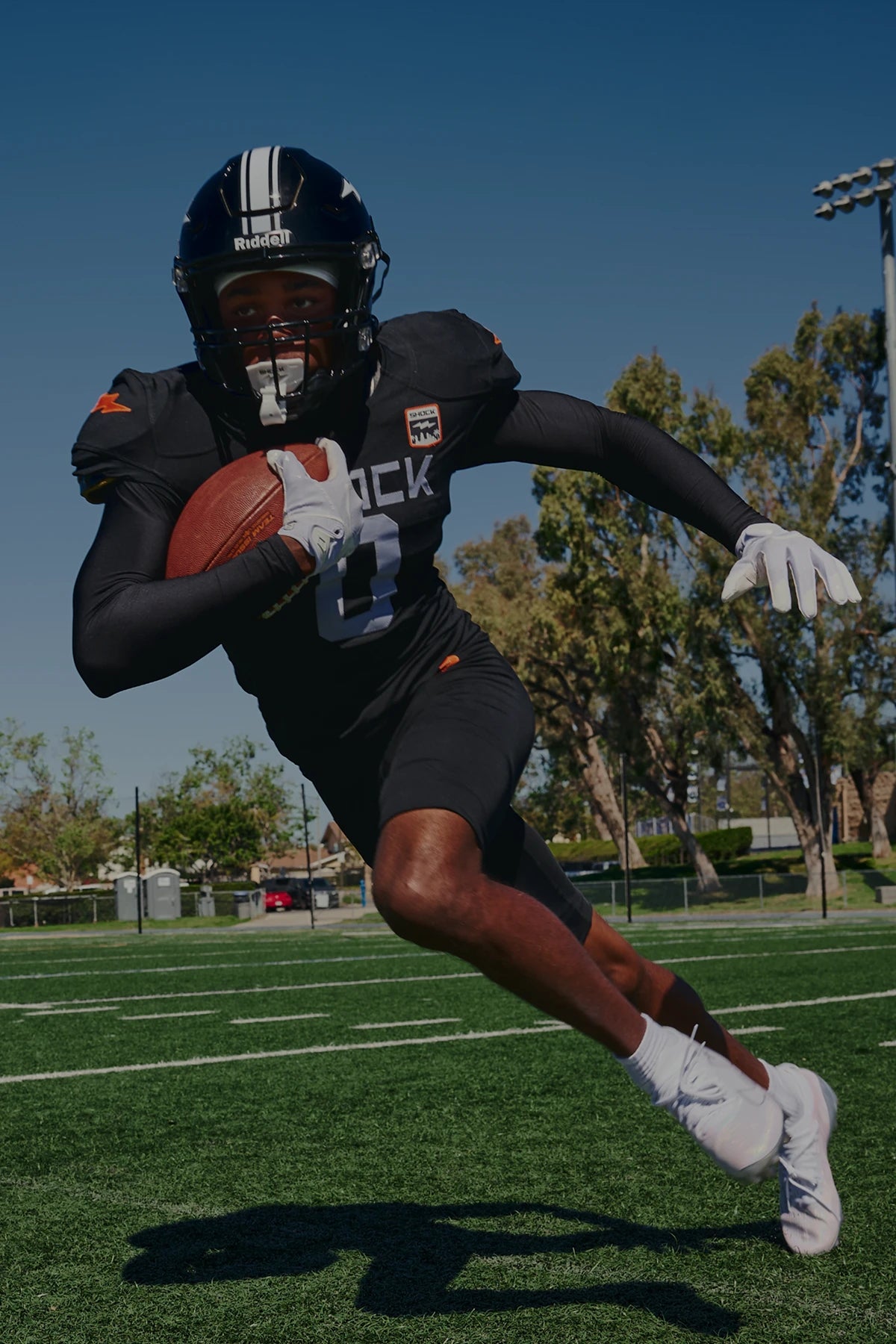 A football player in a black uniform with white accents runs across a green turf field, holding a football under his arm. Trees and a blue sky are visible in the background.