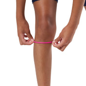 A close-up of a person's leg and hands measuring a bright pink athletic band around the knee, set against a plain white background.