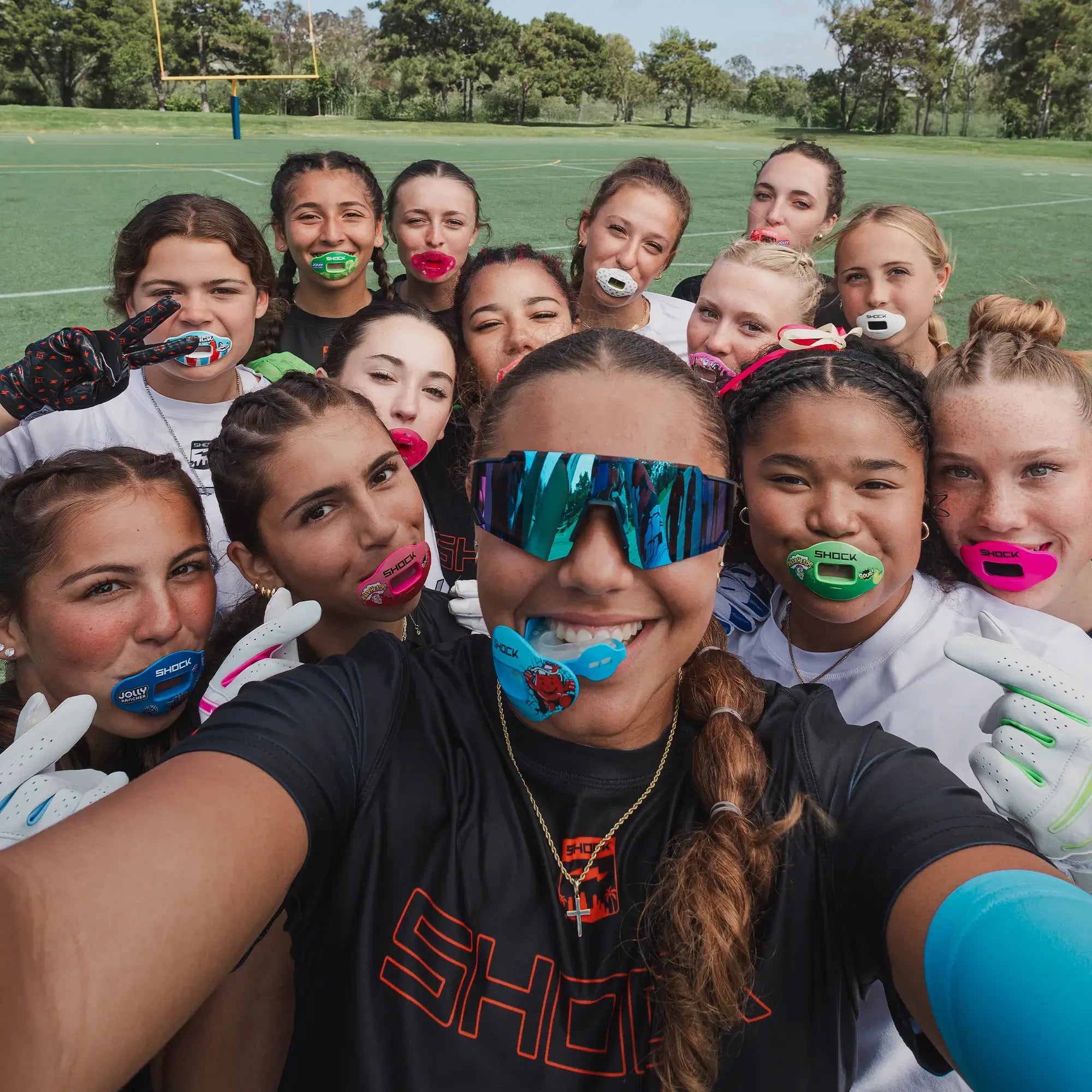 Group of young women posing for a selfie on a sports field, wearing colorful mouthguards.