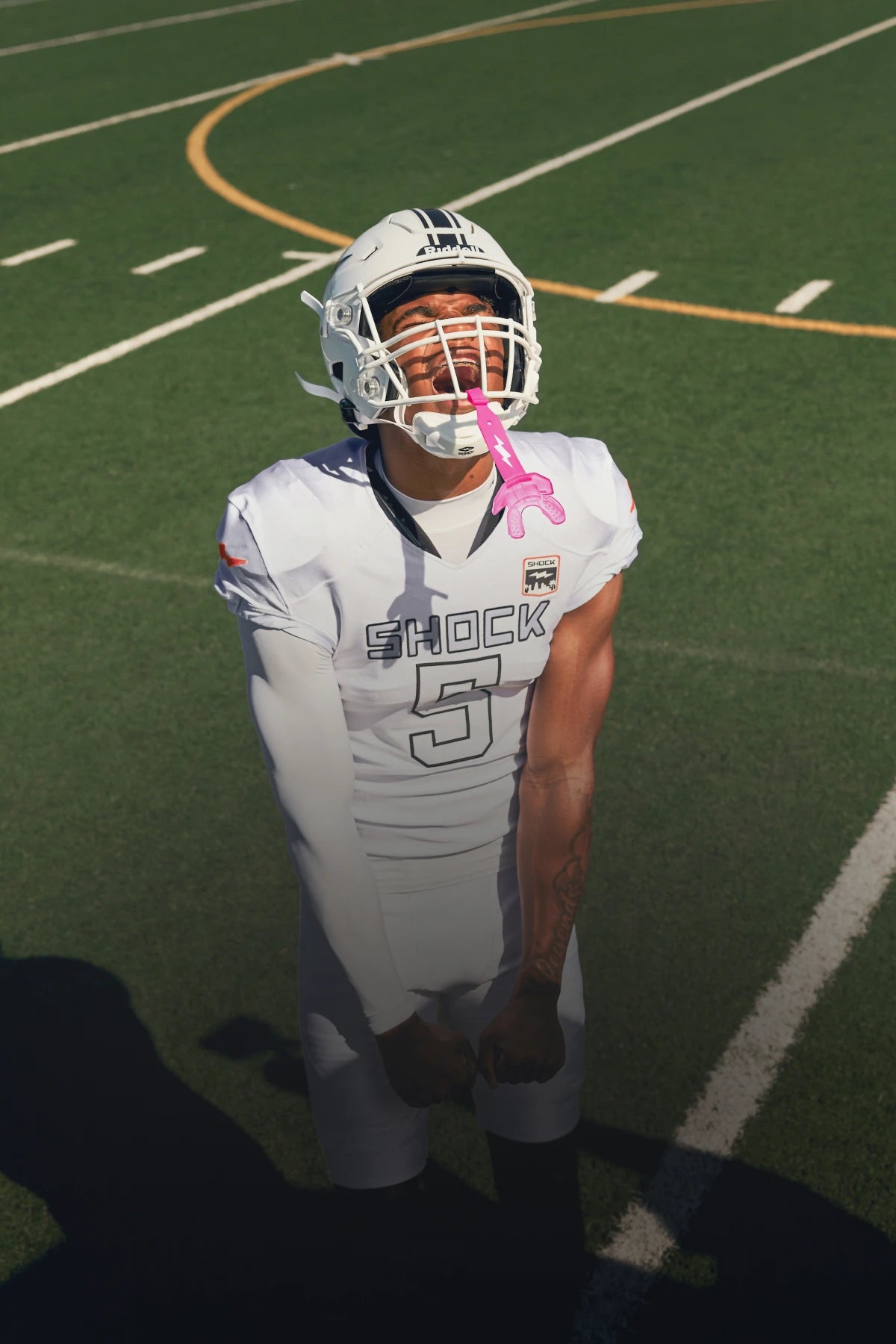 Football player in a white Shock jersey yells skyward on the field, a pink Shock Doctor Thunder Bolt lip guard hanging from his helmet.