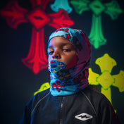 Person wearing a colorful blue sheisty ski mask with abstract designs against a dark background with multicolored abstract shapes.
