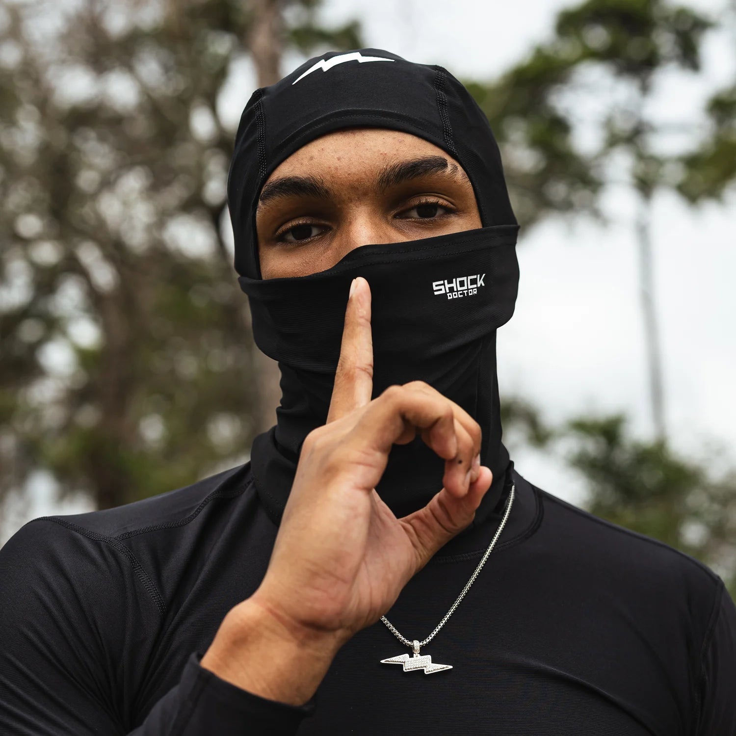 A man wearing a black full-face balaclava mask with a white 'SHOCK DOCTOR' logo, posing with his finger to his lips in an outdoor setting with blurred trees in the background.